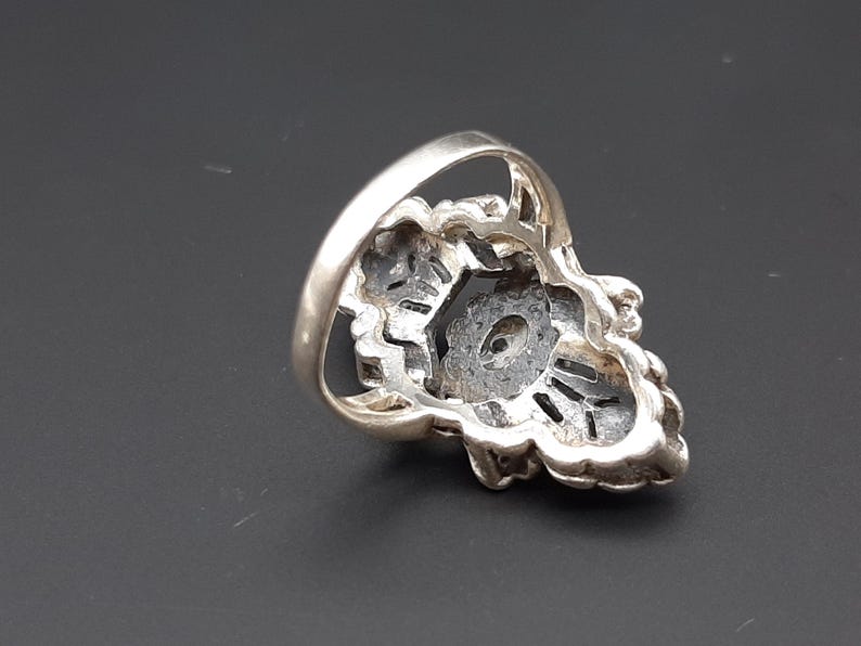 May include: Silver-toned ring with a floral design. The ring features a central dark stone surrounded by intricate metalwork. The band is smooth and rounded.