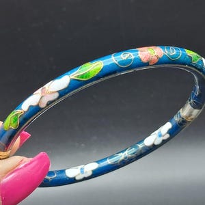 May include: A blue enamel bangle bracelet with a floral design. The bracelet features white, pink, and green flowers with gold accents. The background is a dark blue color.