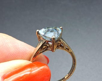Può includere: Un anello in oro con una pietra preziosa azzurra dal taglio triangolare, fissata da una montatura a quattro griffe. La fascia presenta un intricato design in filigrana traforata sui lati, con una finitura metallica e una pietra sfaccettata.
