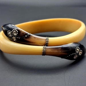 May include: A two-piece bangle bracelet set. The bracelets are a light yellow color with dark brown accents. The ends of the bracelets have a dark brown color with small, clear, round embellishments. The bracelets are connected with a small, silver-colored chain.