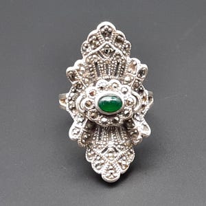 May include: A silver-toned ring with an ornate, Art Deco-inspired design. The ring features a central oval-shaped green stone, surrounded by smaller, sparkling accents. The ring's intricate details and symmetrical pattern create a vintage aesthetic.