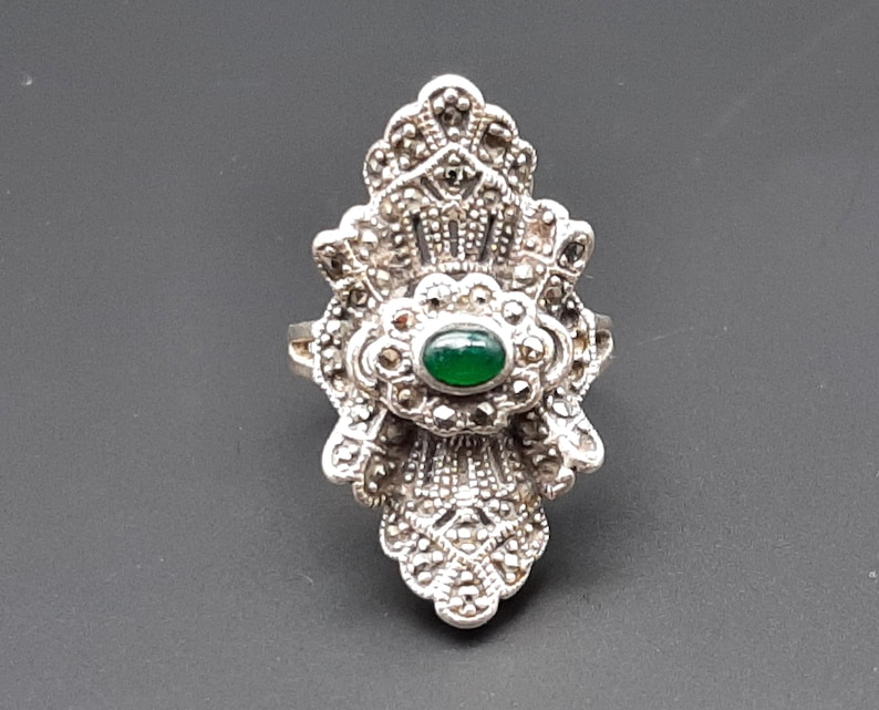 May include: A silver-toned ring with an ornate, Art Deco-inspired design. The ring features a central oval-shaped green stone, surrounded by smaller, sparkling accents. The ring's intricate details and symmetrical pattern create a vintage aesthetic.