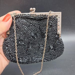 Könnte beinhalten: Eine schwarze Handtasche, die vollständig mit kleinen schwarzen Glasperlen in einem floralen Muster bestickt ist. Die Tasche verfügt über einen silberfarbenen Metallrahmen mit einem Schnappverschluss und eine feine silberne Metallkette.