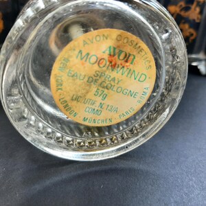 May include: A clear glass bottle with a yellow label that reads "Avon Cosmetics Moonwind Spray Eau de Cologne 57g Lic. Utif. N. 13/A Como Munchen Paris Roma New York London".
