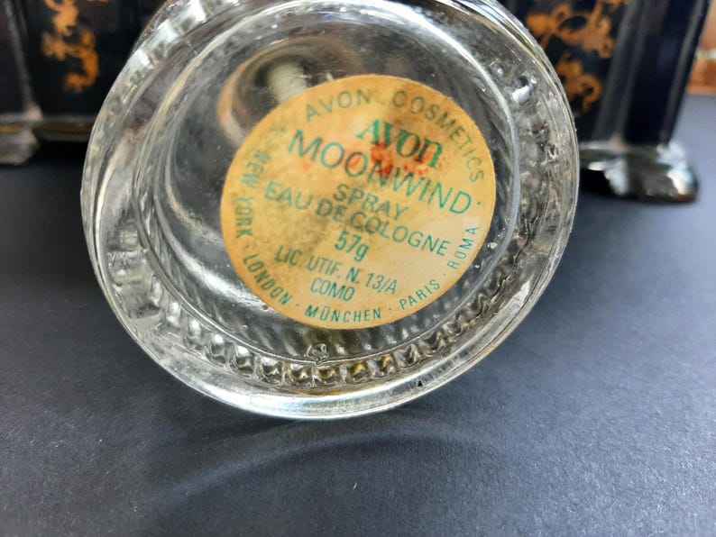 May include: A clear glass bottle with a yellow label that reads "Avon Cosmetics Moonwind Spray Eau de Cologne 57g Lic. Utif. N. 13/A Como Munchen Paris Roma New York London".