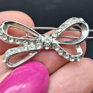 May include: A silver-toned metal hair clip shaped like a bow, featuring a cutout design embellished with small, clear, sparkling rhinestones along the entire front surface of the bow and the central knot, with a standard metal snap-style closure.