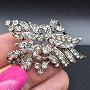 Op de afbeelding: Een zilverkleurige broche met een bloemachtig ontwerp, voorzien van meerdere heldere, gefacetteerde strass-steentjes in het metalen frame. Het sieraad heeft een gebogen, lintachtige structuur met kleine groepjes ronde steentjes aan de randen.