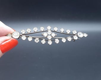 Könnte beinhalten: Silberfarbene Brosche mit symmetrischem Design. Die Brosche hat klare, runde Edelsteine, die entlang einer gebogenen, länglichen Form angeordnet sind. Die Edelsteine sind gleichmäßig verteilt und erzeugen einen funkelnden Effekt. Die Brosche wird von einer Hand mit rotem Nagellack gehalten.
