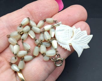 May include: A necklace featuring a series of small, teardrop-shaped, off-white mother-of-pearl beads linked together with thin metal wire. The necklace includes a carved mother-of-pearl fish pendant and a circular metal spring ring clasp.