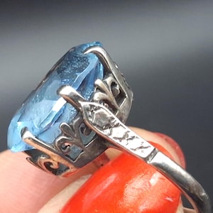Pode incluir: Um anel de prata com uma grande pedra preciosa azul-clara facetada, mantida numa cravação de metal decorativa com detalhes recortados intrincados ao longo da galeria e um padrão gravado texturizado no aro do anel.