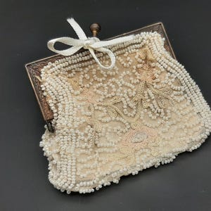 Puede incluir: Un antiguo monedero de cuentas con un marco de metal y un lazo blanco. El monedero está cubierto de pequeñas cuentas blanquecinas con un diseño floral en rosa pálido y beige. El marco tiene un aspecto decorativo y envejecido.