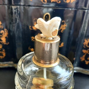 May include: A clear glass bottle with a gold-colored top and a white bird-shaped stopper. The bottle is sitting on a black surface with gold designs.