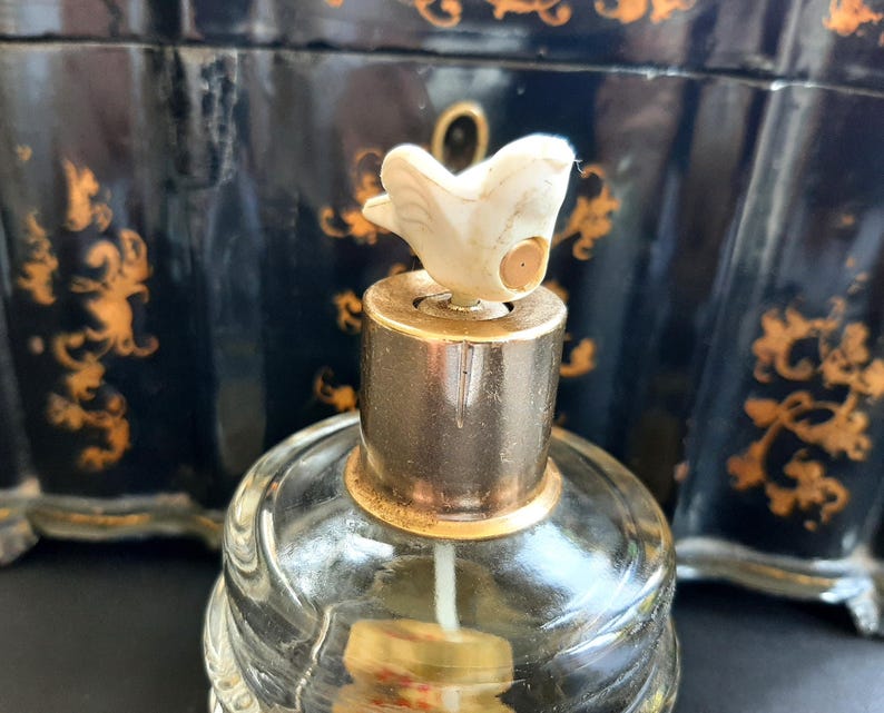 May include: A clear glass bottle with a gold-colored top and a white bird-shaped stopper. The bottle is sitting on a black surface with gold designs.