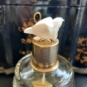 May include: A vintage glass perfume bottle with a gold top and a white bird-shaped stopper. The bottle is sitting on a black and gold surface.