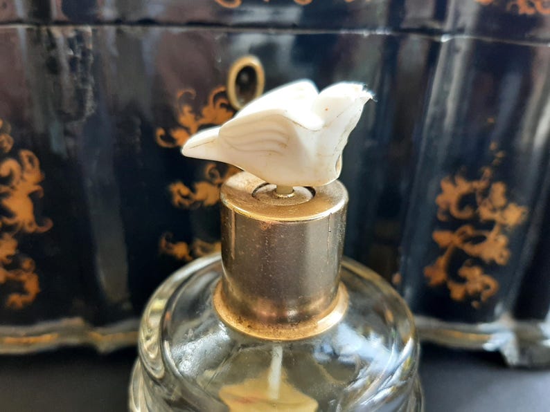 May include: A vintage glass perfume bottle with a gold top and a white bird-shaped stopper. The bottle is sitting on a black and gold surface.