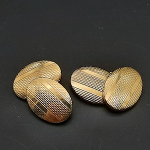 May include: Four gold-coloured oval cufflinks with textured diagonal and parallel lines. The cufflinks are arranged in two pairs, each with a matching design. The background is a solid black.