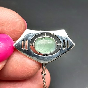 May include: A silver pendant with a light green oval-shaped stone at the center. The pendant has a geometric design with a chain attached. The background is a dark gray.