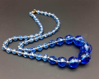 Op de afbeelding: Een blauwe kralenketting met facetkralen. De ketting heeft een mix van licht- en donkerblauwe kralen, geregen met zilverkleurig metaal. De sluiting is goudkleurig. De ketting wordt gepresenteerd op een donkere ondergrond.