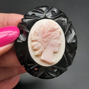 May include: Oval black cameo brooch featuring a profile of a woman with a pink face and hair, set against a white background. The black frame has a decorative, carved design. A classic piece of jewelry.