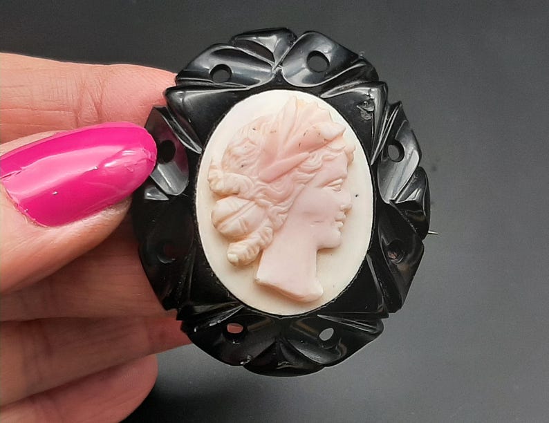 May include: Oval black cameo brooch featuring a profile of a woman with a pink face and hair, set against a white background. The black frame has a decorative, carved design. A classic piece of jewelry.