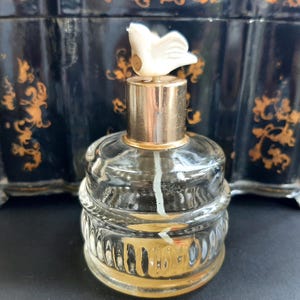 May include: A clear glass bottle with a gold top and a white bird-shaped stopper. The bottle has a textured design and is filled with a clear liquid.