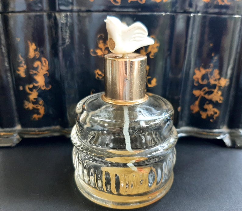 May include: A clear glass bottle with a gold top and a white bird-shaped stopper. The bottle has a textured design and is filled with a clear liquid.