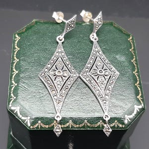 Puede incluir: Un par de pendientes colgantes plateados. Cada pendiente presenta un diseño en forma de diamante con un motivo floral central y está incrustado con pequeñas piedras brillantes. Los pendientes se exhiben en una caja de joyería verde.