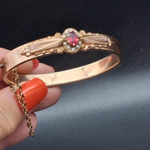 Può includere: Un bracciale rigido incernierato color oro con una pietra preziosa rossa di forma ovale al centro, circondata da piccole perle bianche. La fascia metallica presenta dettagli decorativi in rilievo su entrambi i lati e include una catenella di sicurezza.