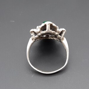 May include: Silver ring with a detailed floral design and a small, round, green gemstone at the center. The ring's band is smooth and polished, and the setting has intricate detailing.