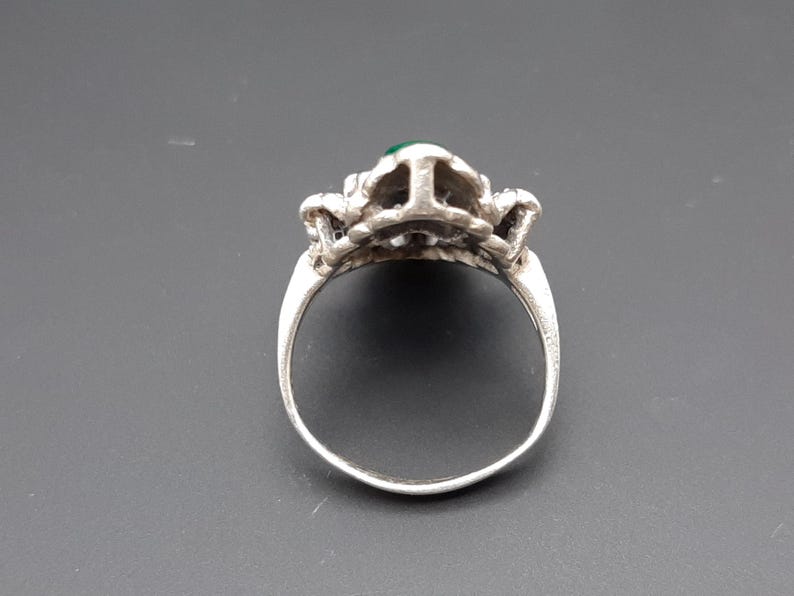 May include: Silver ring with a detailed floral design and a small, round, green gemstone at the center. The ring's band is smooth and polished, and the setting has intricate detailing.