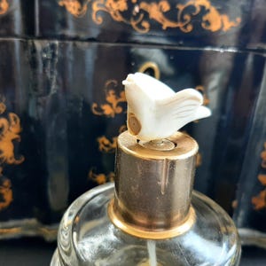 May include: A vintage glass bottle with a gold top and a white bird-shaped stopper. The bottle is sitting on a black surface with gold accents.