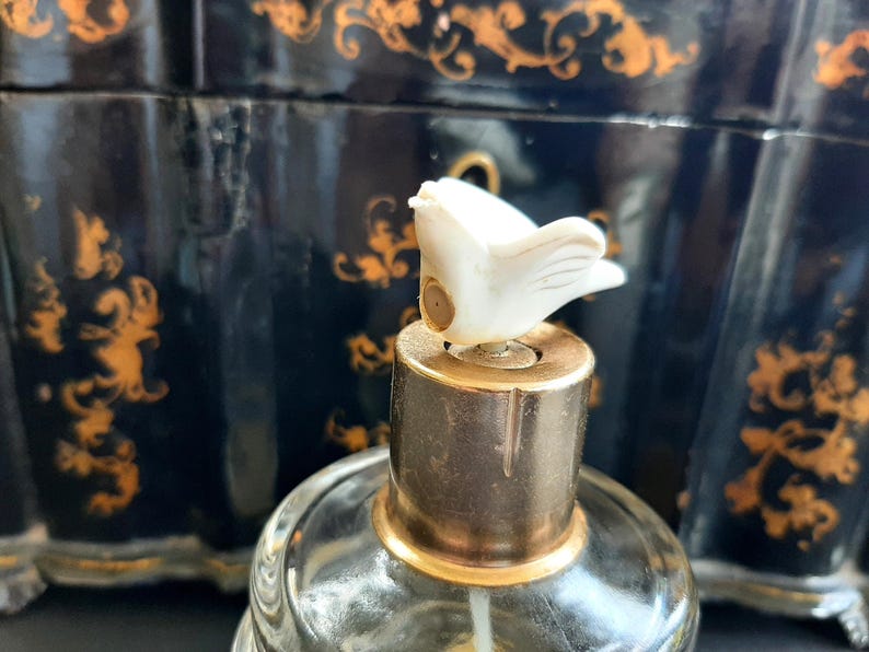 May include: A vintage glass bottle with a gold top and a white bird-shaped stopper. The bottle is sitting on a black surface with gold accents.