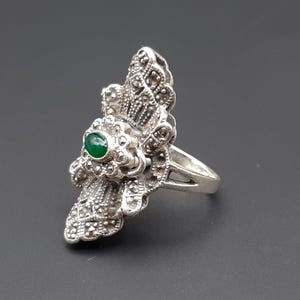May include: A silver-toned ring with an ornate design. The ring features a central green gemstone and is embellished with smaller, sparkling accents. The ring's design is detailed and intricate, with a vintage or antique aesthetic.