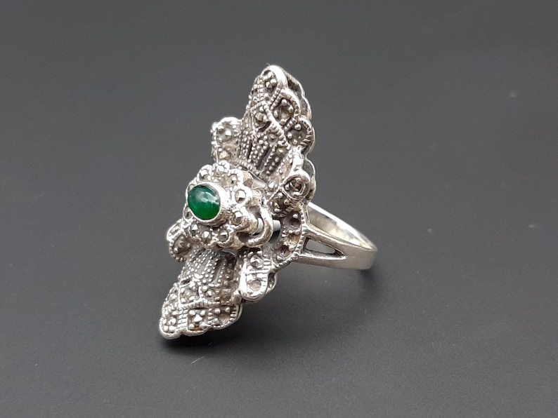 May include: A silver-toned ring with an ornate design. The ring features a central green gemstone and is embellished with smaller, sparkling accents. The ring's design is detailed and intricate, with a vintage or antique aesthetic.