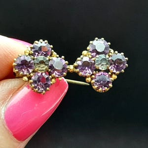 Op de afbeelding: Een vintage goudkleurige broche met twee bloemvormige clusters. Elke cluster is versierd met paarse en groene edelstenen. De broche wordt vastgehouden door een hand met roze nagellak tegen een zwarte achtergrond.