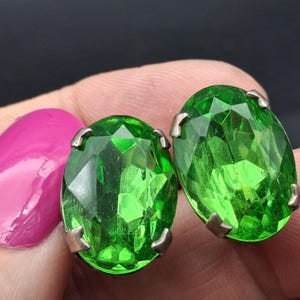 May include: A pair of oval-shaped, faceted bright green glass stones set in silver-toned metal prongs. The stones feature a brilliant cut with multiple reflective surfaces, creating a vibrant emerald-like appearance in this jewelry component.