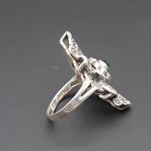 May include: Silver-toned ring with a detailed, ornate design. The ring features a central green stone and is embellished with smaller, sparkling accents. The ring's design is intricate and eye-catching, showcasing a vintage or antique aesthetic.