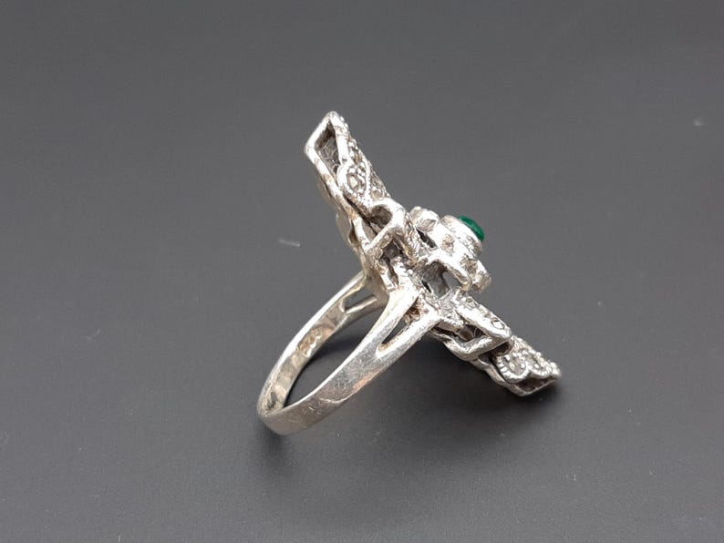 May include: Silver-toned ring with a detailed, ornate design. The ring features a central green stone and is embellished with smaller, sparkling accents. The ring's design is intricate and eye-catching, showcasing a vintage or antique aesthetic.
