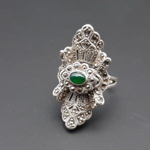 May include: Silver-toned ring with a central oval green stone, surrounded by intricate marcasite detailing. The ring has a decorative, fan-like design, and is set against a gray background. A vintage-style piece of jewelry.
