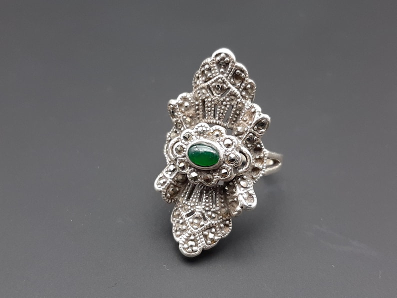 May include: Silver-toned ring with a central oval green stone, surrounded by intricate marcasite detailing. The ring has a decorative, fan-like design, and is set against a gray background. A vintage-style piece of jewelry.