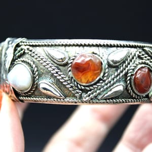 Könnte beinhalten: Ein silbernes Armband mit komplizierten Designs und mehreren roten und orangefarbenen Edelsteinen. Das Armband hat auf einer Seite einen perlmuttartigen Stein.