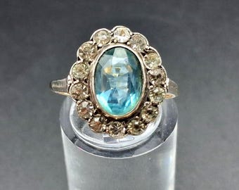 Può includere: Un anello color oro con una grande pietra centrale ovale di colore azzurro, circondata da un alone di dodici pietre più piccole, trasparenti e dal taglio rotondo. La fascia in metallo presenta un design testurizzato e a coste sui lati.