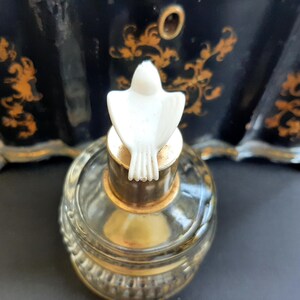 May include: A clear glass bottle with a gold top and a white dove-shaped stopper. The bottle is sitting on a black surface.