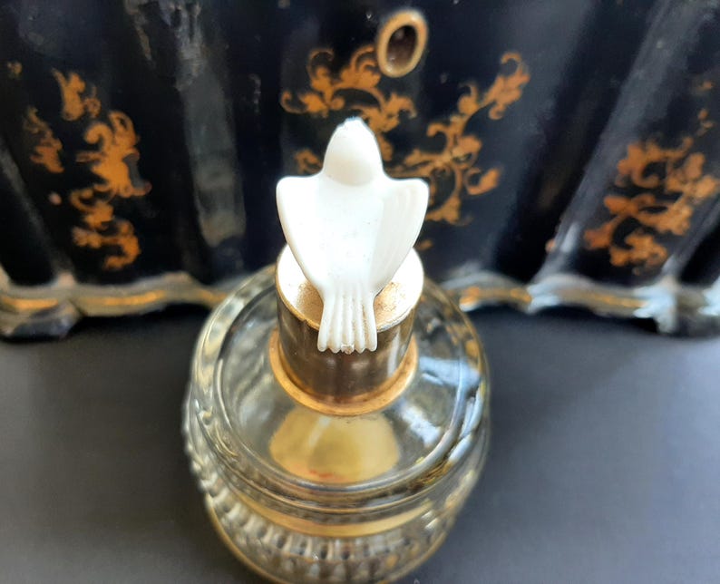 May include: A clear glass bottle with a gold top and a white dove-shaped stopper. The bottle is sitting on a black surface.