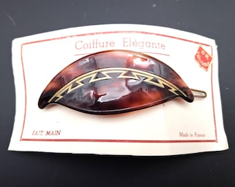 Puede incluir: Una pinza para el pelo en forma de media luna con un patrón de carey marrón y una incrustación geométrica dorada en zigzag. La pinza está sujeta a una tarjeta blanca con el texto Coiffure Elégante, Fait Main y Made in France.