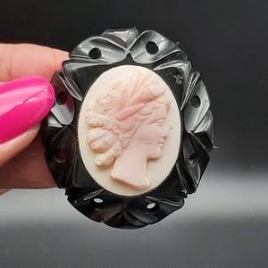 May include: Oval black cameo brooch featuring a profile of a woman with a pink face and hair, set against a cream background. The brooch has a decorative black frame with cut-out details. A classic piece of jewelry.