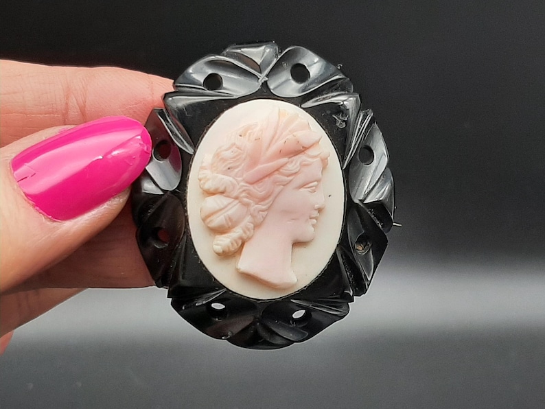 May include: Oval black cameo brooch featuring a profile of a woman with a pink face and hair, set against a cream background. The brooch has a decorative black frame with cut-out details. A classic piece of jewelry.