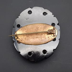 May include: A vintage black brooch with a decorative, irregular edge. The brooch features a gold-toned pin and a brass-colored oval backing. The brooch has several circular cutouts around the edge.