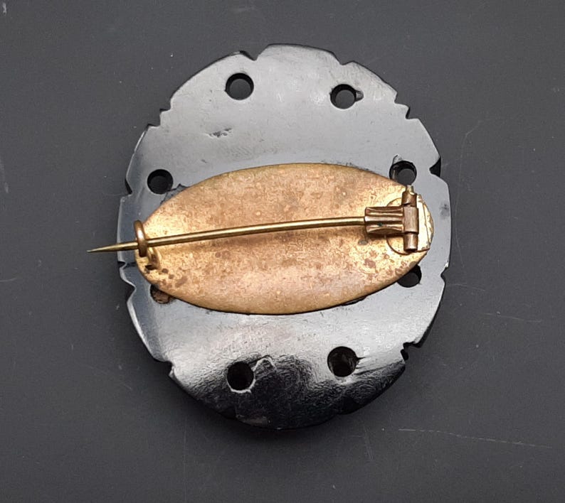 May include: A vintage black brooch with a decorative, irregular edge. The brooch features a gold-toned pin and a brass-colored oval backing. The brooch has several circular cutouts around the edge.