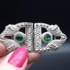 May include: A silver-toned Art Deco buckle with a hinged design. The buckle features two emerald green stones and several clear, round stones. The edges are detailed with small, round accents. The background is a dark, solid color.